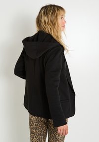 Chaqueta con capucha negra de diseño ajustado, con una tela texturizada y dos bolsillos laterales, combinada con pantalones de estampado de leopardo.