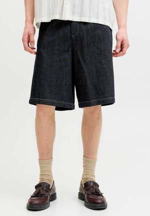 Muž nosiaci tmavé rifľové šortky po kolená, béžové ponožky, hnedé kožené mokasíny s strapcami a bielu košeľu s tenkými vertikálnymi pásikmi.