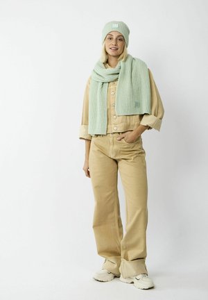 Pale green knitted beanie and matching scarf with ribbed texture. Model wears a tan corduroy jacket and pants, with white sneakers.