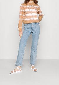 Hnědá batikovaná krátká mikina, světle modré džíny s rovnými nohavicemi a bílé sandály s pásky kolem kotníku. Hladké textury, ležérní střih, minimalistický design.