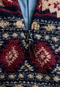 Cremallera negra en una chaqueta de forro polar con un patrón geométrico texturizado en rojo, azul marino y crema sobre una tela azul clara debajo.