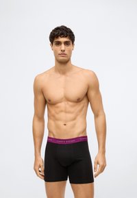 Homem com cabelo curto e encaracolado, usando boxers pretos com uma cintura roxa, de pé contra um fundo branco liso.