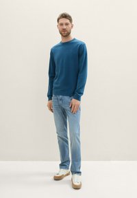 Pulover albastru cu guler rotund, dintr-un tricot ușor, asortat cu blugi decolorati, albaștri și adidași albi cu accente maro.