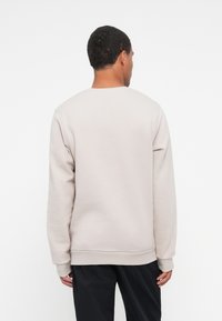 Sudadera de color beige claro con cuello redondo clásico, mangas largas y puños de canalé. Textura suave y diseño minimalista, vista desde atrás.