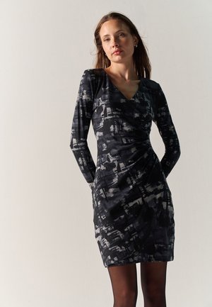Mujer que lleva un vestido de manga larga con estampado abstracto en negro y gris, de pie contra un fondo claro.