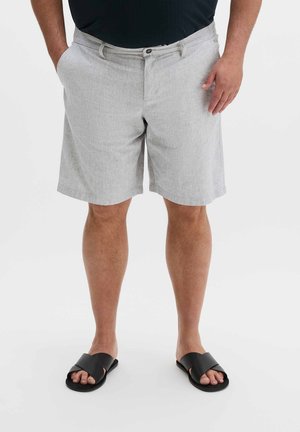 Mann trägt hellgraue knielange Shorts, schwarzes Hemd und schwarze Slipper, steht mit einer Hand in der Tasche.