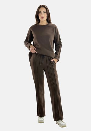 Vrouw draagt een bruine bijpassende sweatshirt en broek met verticale strepen, staat met één hand in de zak, witte sneakers, neutrale achtergrond.