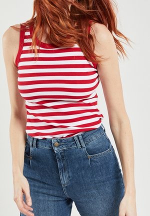 Punahiuksinen nainen pukeutuu punavalkoraidalliseen hihattomaan toppiin ja korkeavyötäröisiin sinisiin farkkuihin yksiväristä taustaa vasten.