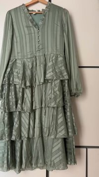 Πράσινο, στρωτό φόρεμα με μακριά μανίκια, κατακόρυφες ρίγες και V-λαιμόκοψη. Διαθέτει κουμπιά κατά μήκος του μπροστινού μέρους και ρέουσα κρόσσια. Φτιαγμένο από ελαφρύ ύφασμα.