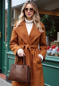 Bruine wollen jas met een strikceintuur, witte turtleneck en bruine leren handtas. Ronde zonnebril voegt detail toe aan de outfit. Groene achtergrond.