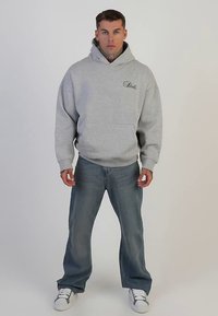 Hanorac gri deschis cu buzunar frontal, glugă cu șnur și logo brodat. Asortat cu blugi albaștri largi și adidași albi.