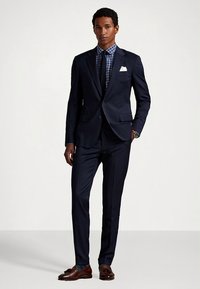 Marineblå dress med smal passform, kombinert med en rutete skjorte og slips. Har et hvitt lommetørkle og brune skinnloafers.