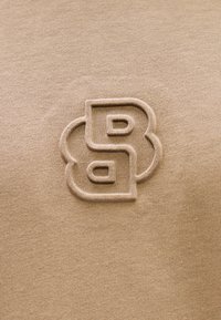 Μπεζ ύφασμα με λεία υφή, που έχει κεντημένο λογότυπο σε αντίθετο καφέ χρώμα, κεντραρισμένο στο υλικό.