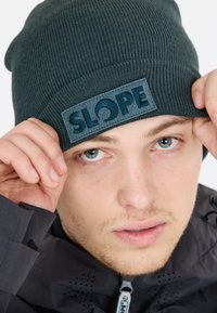 Mörkgrön stickad beanie med vikbar mudd, med en blå texturerad patch märkt "SLOPE." Slät textur och tight passform.