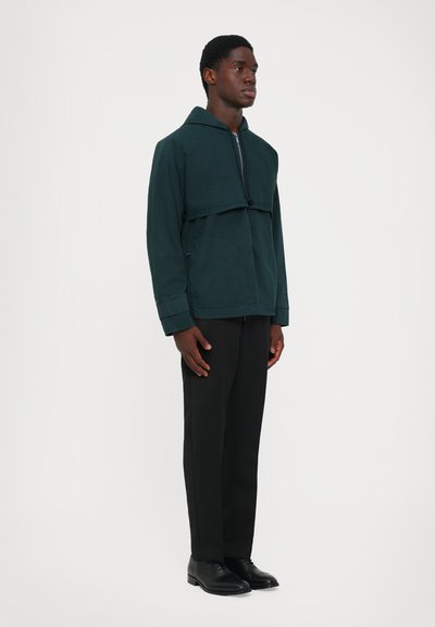 Άνδρας που φοράει σκούρο πράσινο φερμουάρ παντελόνι με φαρδύ γιακά, μαύρο παντελόνι και μαύρα κοστούμια, που στέκεται μπροστά από έναν απλό φωτεινό φόντο.