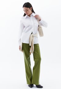 Camicia bianca a maniche lunghe, pantaloni verdi a palazzo, maglione beige legato in vita, scarpe nere e borsa a tracolla color crema. Tessuto liscio, design strutturato.