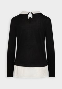 Svart långärmad topp med vit lager längst ner och vit krage, med ett nyckelhål bak och kontrasterande tyg vid halsringningen.