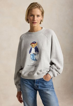 Giovane donna con felpa grigia con grafica orso, vestita in abiti casual e jeans blu, in posa con una mano in tasca.