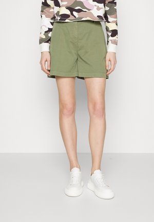 Persona que lleva pantalones cortos verde oliva, una camisa de camuflaje de manga larga y zapatillas blancas, de pie contra un fondo liso.