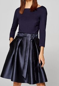 Kvinna klädd i en marinblå topp med trekvartsärmar och en högmidjad svart satin A-linjefald i kombination med ett brett bälte knutet framtill.