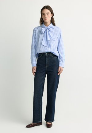 Junge Frau trägt eine blau-weiß karierte Bluse mit großer Schleife und dunkle, hochtaillierte Jeans mit geradem Bein, steht vor einem schlichten Hintergrund.