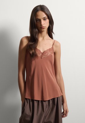 Junge Frau mit langen dunklen Haaren, die eine rostfarbene Spitze-trimmed Camisole und lockere braune Hose vor einem schlichten hellen Hintergrund trägt.
