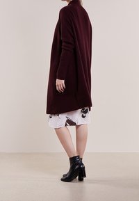 Cardigan lungo bordeaux con una vestibilità rilassata, texture a coste e tasche laterali, abbinato a una gonna bianca con motivo floreale e stivali neri alla caviglia.