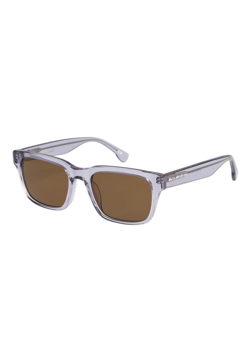 Gafas de sol de plástico transparente con montura cuadrada, lentes de color marrón, y brazos curvados y elegantes. Presentan un branding mínimo en el lateral.