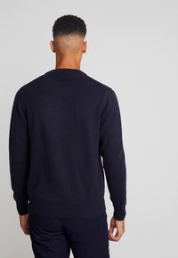 Maglione a maglia blu navy con colletto e polsini a coste, caratterizzato da una texture solida e un orlo aderente, indossato da una persona rivolta di spalle rispetto alla fotocamera.