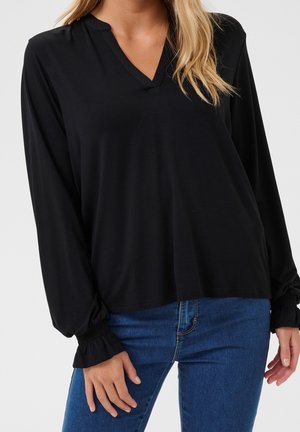 Kvinde iført en sort bluse med lange ærmer og V-hals med rynkede manchetter kombineret med blå jeans mod en ensfarvet baggrund.
