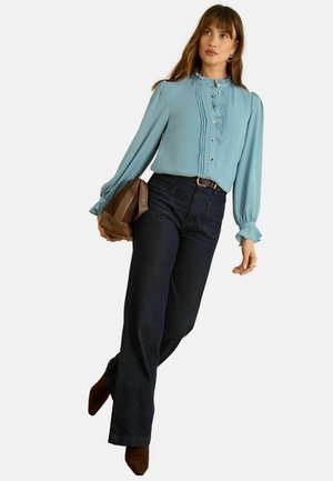 Light blue blouse with ruffled detailing, button closure; paired with dark wide-leg jeans. Model carrying a brown clutch, wearing brown heels.