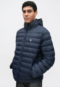 Geacă matlasată de culoare bleumarin cu glugă, având o textură matlasată, închidere cu fermoar și un logo subtil pe piept. Material moale și ușor.