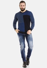 Blaues und marineblaues Sweatshirt mit einem Patchwork-Design und strukturierten Mustern. Getragen mit blauen Jeans und schwarzen Stiefeln.
