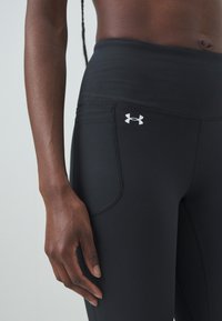 Black athletic leggings featuring a high waistband, smooth texture, side pocket, and a white Under Armour logo on the front.