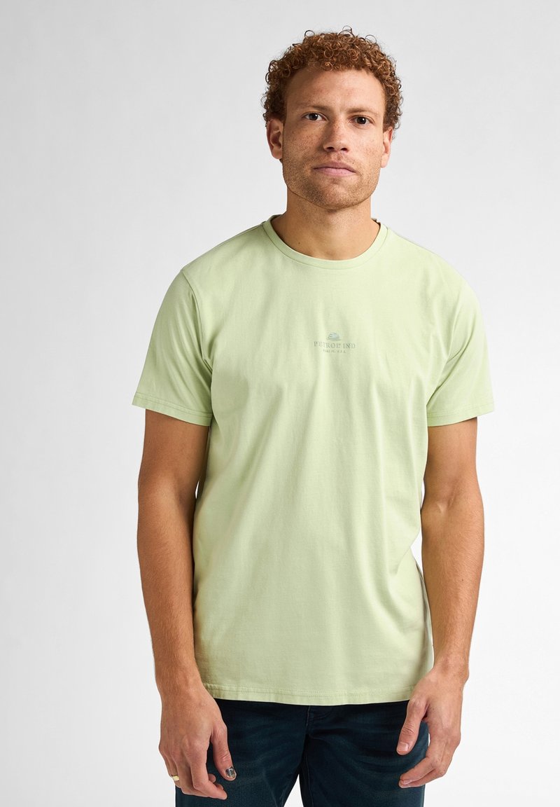 Camiseta de algodón verde claro con cuello redondo y mangas cortas; presenta un pequeño logo impreso en el pecho. Textura suave, ajuste casual.