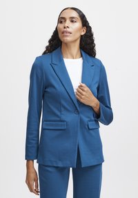 Blauer maßgeschneiderter Blazer mit Reverskragen, einreihiger Knopfverschluss, zwei Fronttaschen und glatter Textur, kombiniert mit einem weißen Oberteil.