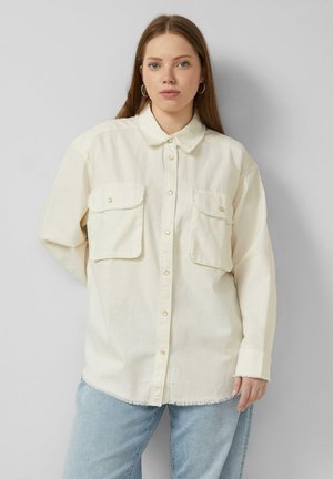 Junge Frau mit langen Haaren, die ein übergroßes cremefarbenes Hemd mit Klapptaschen und hellblaue Jeans vor einem einfarbigen grauen Hintergrund trägt.