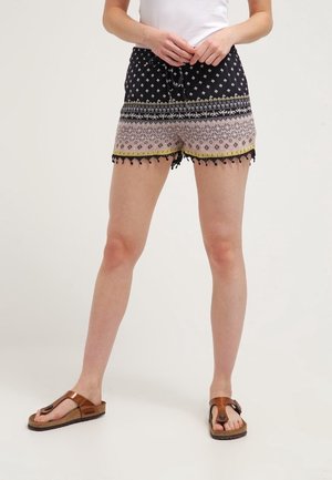 Kvinde iført mønstrede sorte og beige shorts med kvaster, hvid top og brune sandaler, stående med hænderne foldet tæt ved livet.