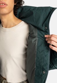 Persoon in een beige geribde crop top trekt een donkergroene jas open om de binnenvoering en de pocket met drukknoop te tonen, tegen een witte achtergrond.