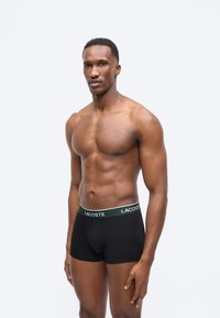 Αθλητικός άνδρας που φοράει μαύρα boxer Lacoste, στέκεται μπροστά σε απλό λευκό φόντο, στραμμένος ελαφρώς αριστερά.