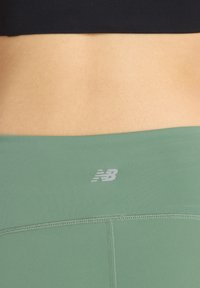Leggings deportivos verdes con una textura suave, costuras sutiles y un logo de New Balance en gris claro en la cintura.