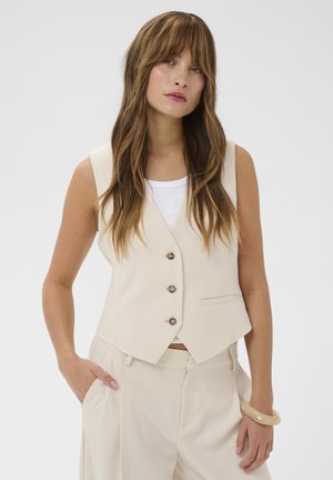 Junge Frau mit langem braunem Haar, die eine beige ärmellose Weste, passend dazu Hosen, ein weißes Oberteil und ein beiges Armband vor weißem Hintergrund trägt.