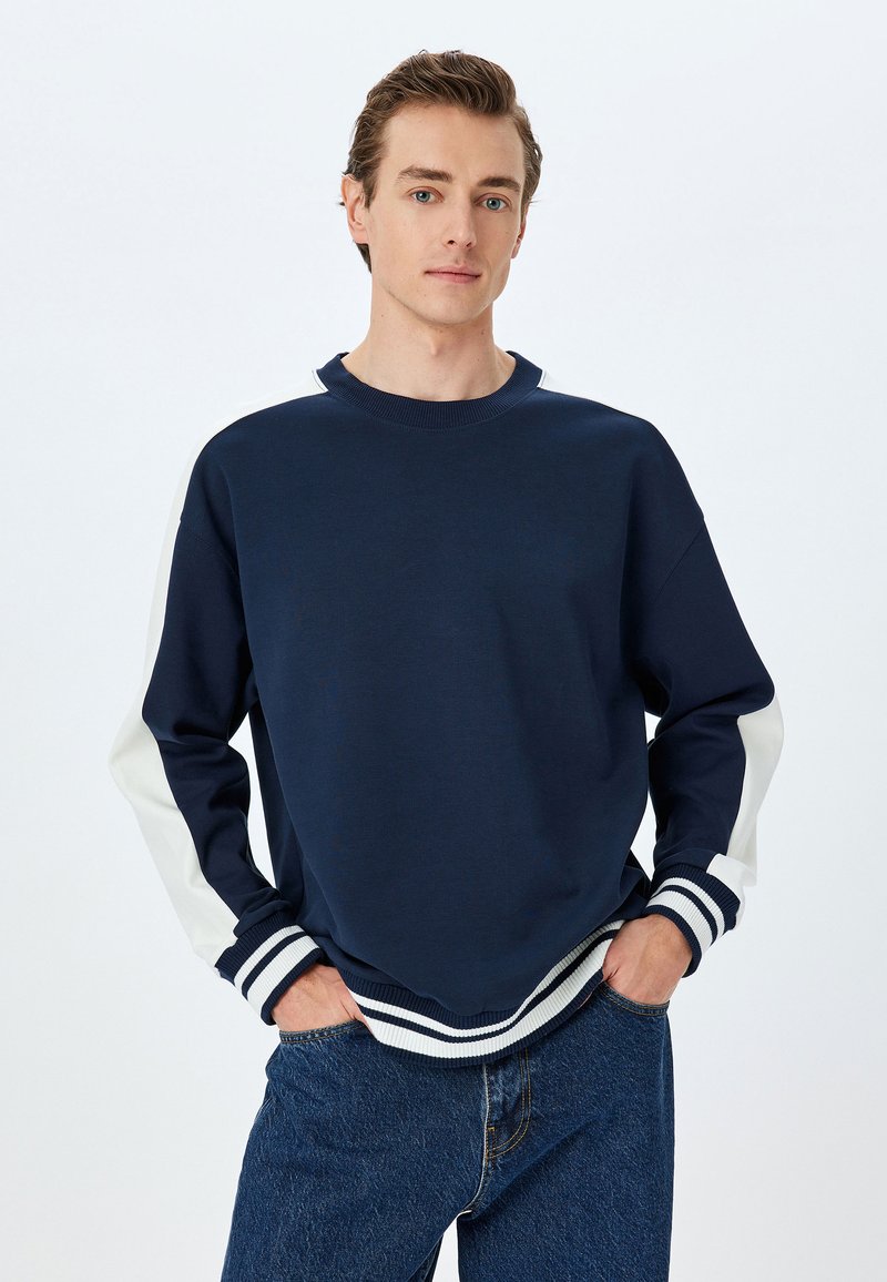 Mörkblå sweatshirt med vita panelaccent på ärmar, ribbade mudd och nederkant, tillverkad av ett mjukt tyg, med en rundad halsringning.