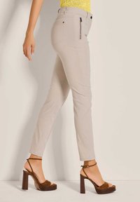 Beige Slim-Fit-Hosen mit einer glatten Textur, ausgestattet mit einer mit Reißverschluss verschließbaren Tasche. Kombiniert mit braunen Riemchensandalen mit auffälliger Sohle.