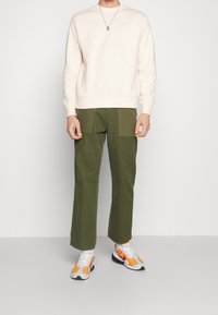 Světle růžová bavlněná mikina, olivově zelené široké kalhoty s kapsami typu cargo a bílo-oranžové tenisky. Viditelný stříbrný náhrdelník.