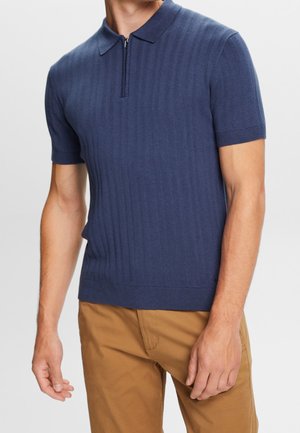 Homme portant un polo à manches courtes bleu marine côtelé avec une fermeture éclair à l'avant et un pantalon de couleur camel, debout devant un fond uni.