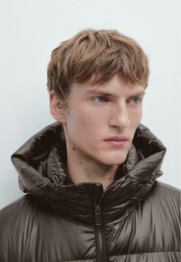 Giacca a vento nera con colletto alto e cappuccio, caratterizzata da una texture lucida e chiusura con cerniera. Il volto del modello è parzialmente visibile.