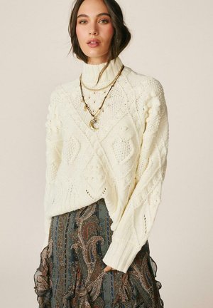 Mujer con suéter de cuello alto de punto tipo cable en color crema, collares en capas y falda oscura con estampado de cachemira, de pie frente a un fondo liso.
