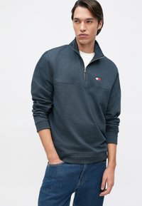 Polaire cinza escuro com colarinho de meia-zíper, punhos canelados e um pequeno patch com logotipo com detalhes em vermelho e azul. Ajuste casual e textura macia.