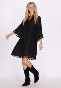 Schwarzes Kleid mit ausgestellter Silhouette, 3/4-Bellärmeln und Troddelbesatz am Saum. Kombiniert mit schwarzen Wildlederstiefeletten.